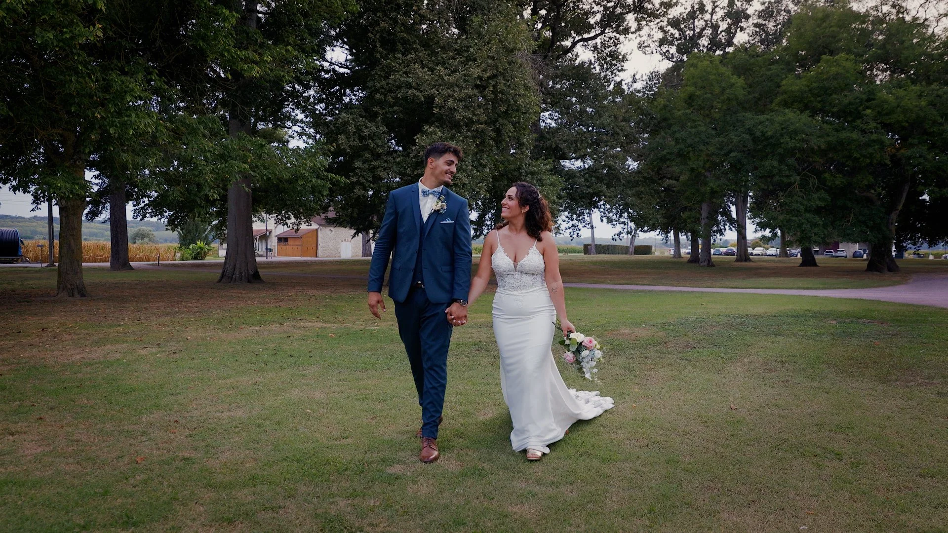 Un couple de mariés marche main dans la main sur l'herbe, entouré d'arbres et de bâtiments en arrière-plan, lors d'une cérémonie ou d'une séance photo en plein air.