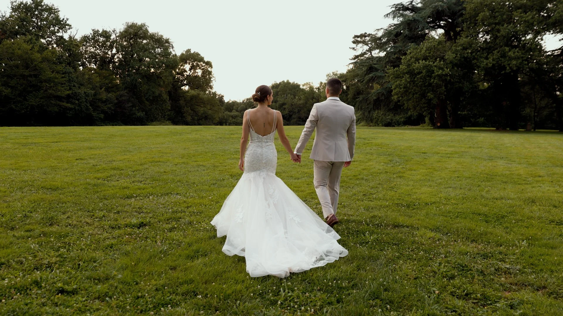 Un couple portant un costume de mariage marche dans un champ vert avec des arbres en arrière-plan, vue de dos.