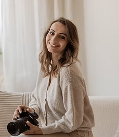 Une jeune femme souriante tenant un appareil photo, assise dans une pièce lumineuse.