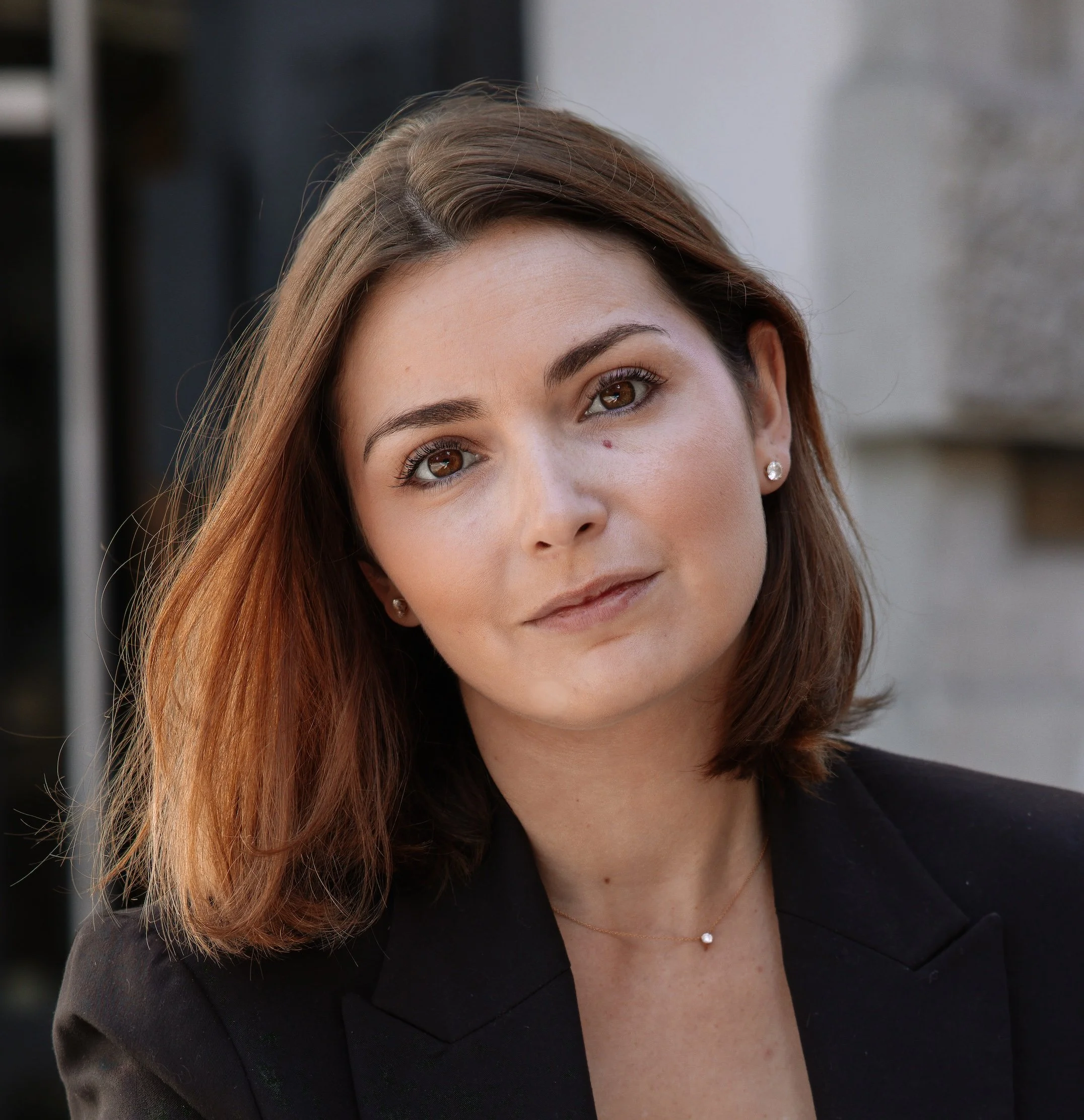 Portrait d'une femme avec des cheveux bruns, portant une veste noire, portant des boucles d'oreilles en diamants et un collier, regardant vers la caméra, en extérieur.