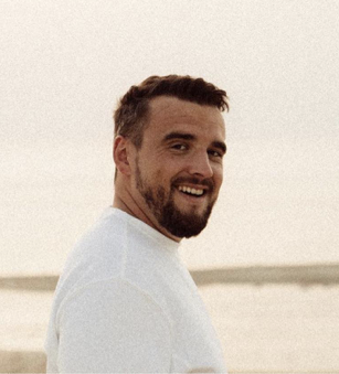 Un homme souriant avec une barbe, portant un t-shirt blanc, à la plage, au coucher du soleil.