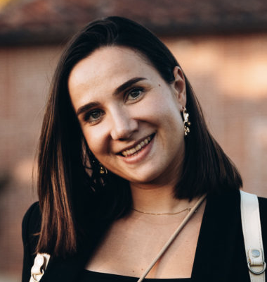 Jeune femme souriante avec des cheveux bruns, portant des boucles d'oreilles et une robe noire, en extérieur.