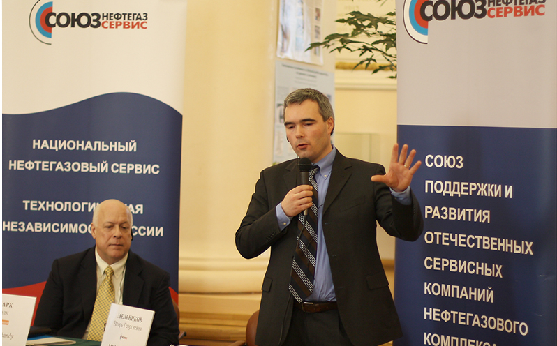 Two men at a conference or meeting with banners in Russian in the background, one standing and speaking into a microphone, the other seated at a table.