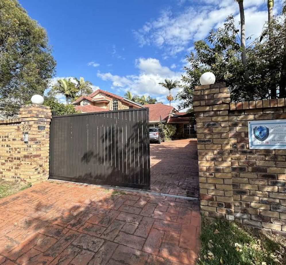 Brick gate and wall at the entrance of a residential property with a red-tiled house and trees in the background under a partly cloudy sky.