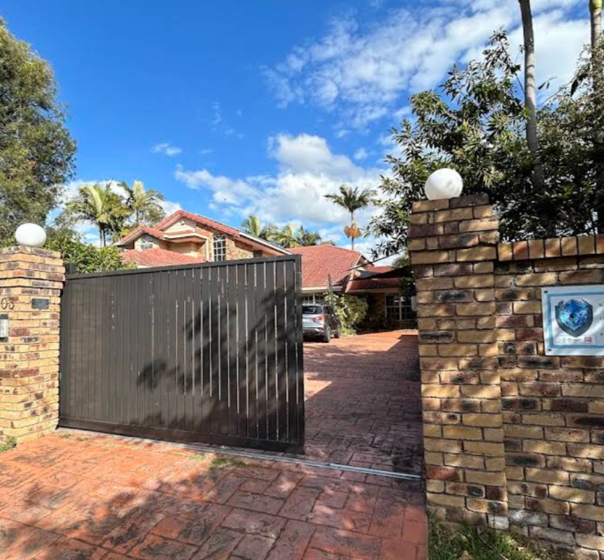 A brick gate with a black sliding door opens into a driveway leading to a house with a red-tiled roof, surrounded by palm trees and greenery under a blue sky with clouds. 603 Musgrave Road, Robertson, Queensland, 4109