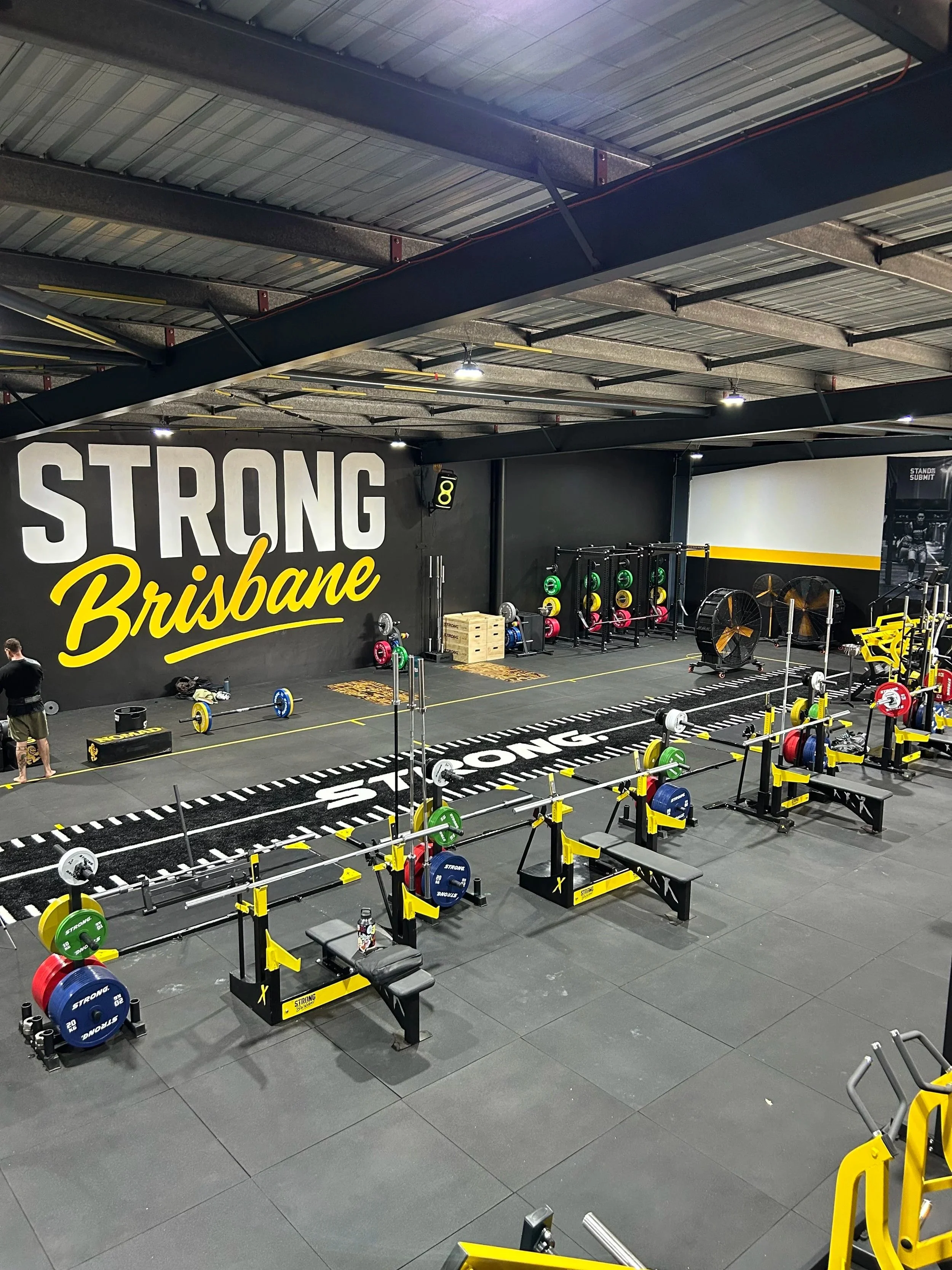 Interior of a gym with strength training equipment, including squat racks with barbells and weight plates, with a large wall mural that says 'Strong Brisbane' in bold letters.