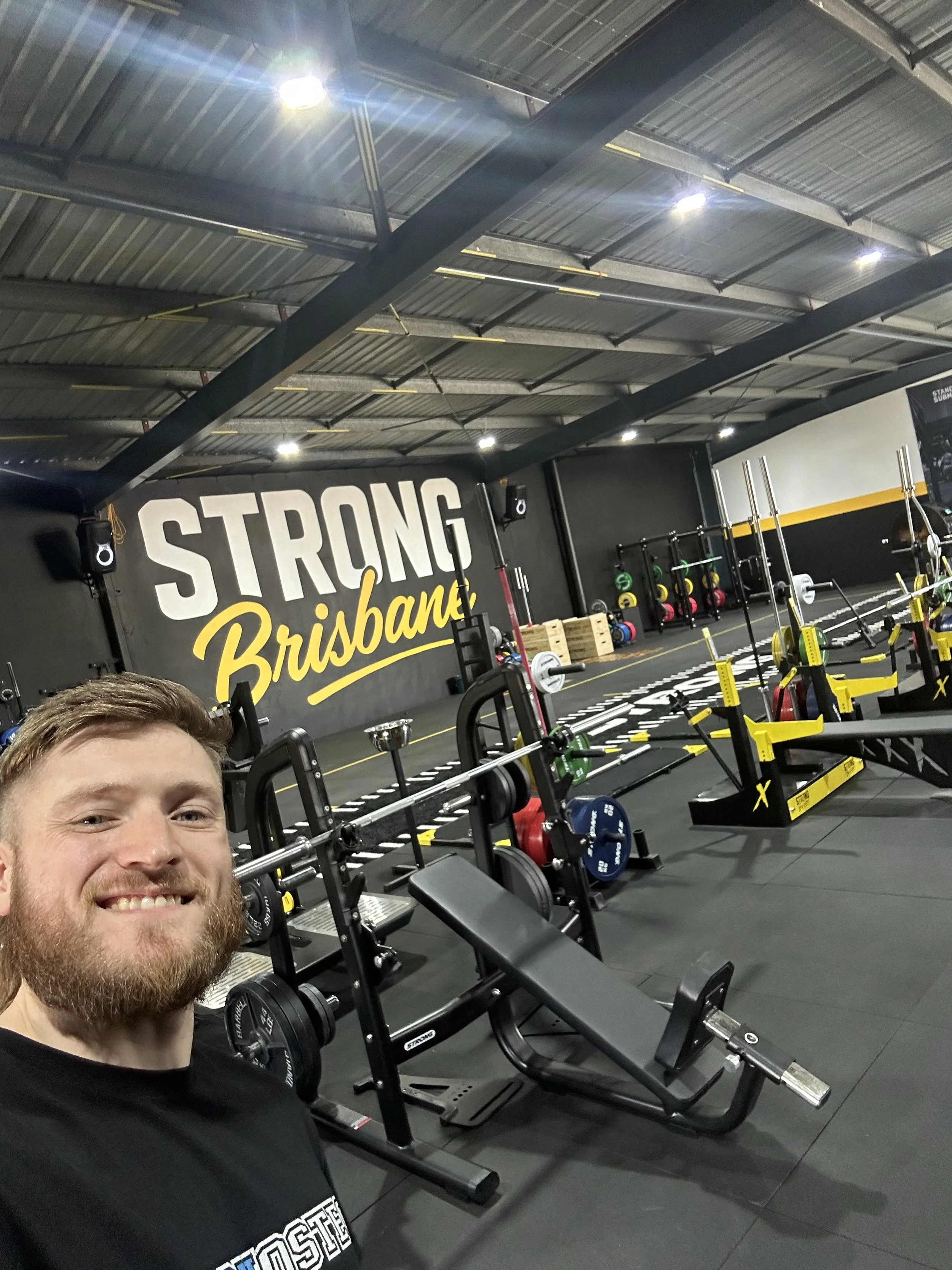 A man, Isaiah Young, taking a selfie inside a gym with weight training equipment, barbells, and a large wall sign that says 'Strong Brisbane'.