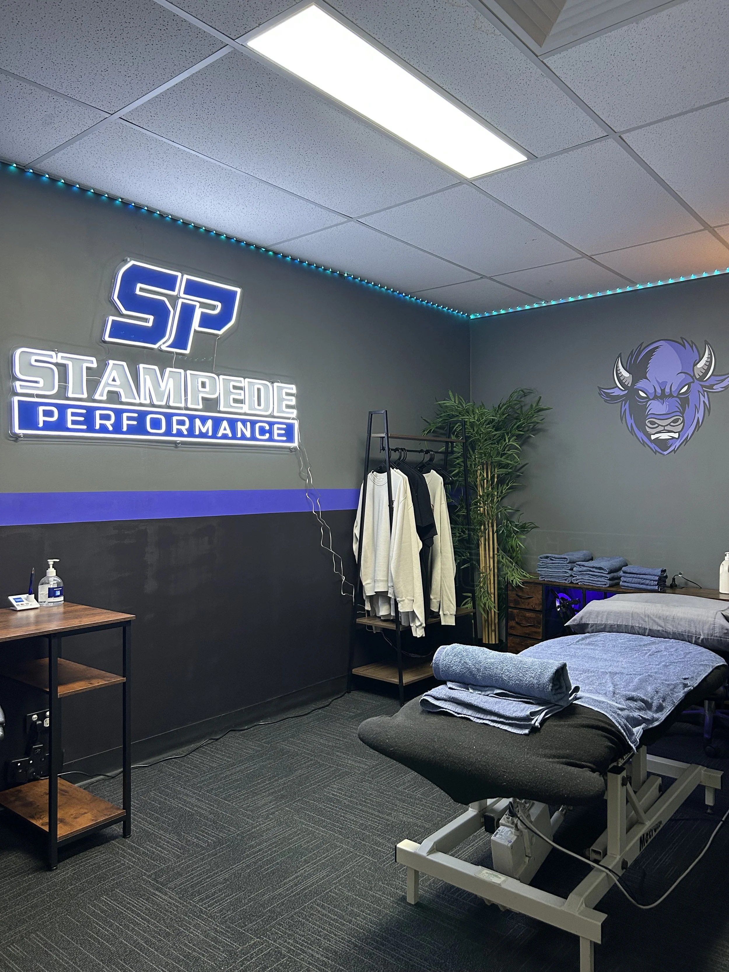 A massage or therapy room with a massage table, rolled towels, clothing rack with sportswear, and neon signs reading "Stampede Performance" and a stylized bison logo, with a leafy plant and folded towels on a wooden dresser.