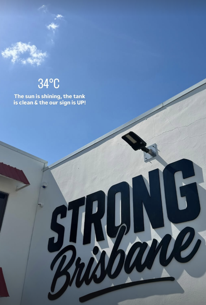 Clear blue sky with a small cloud, a white building with black signage that reads 'STRONG Brisbane', a black outdoor light fixture, and a weather display showing 34°C.
