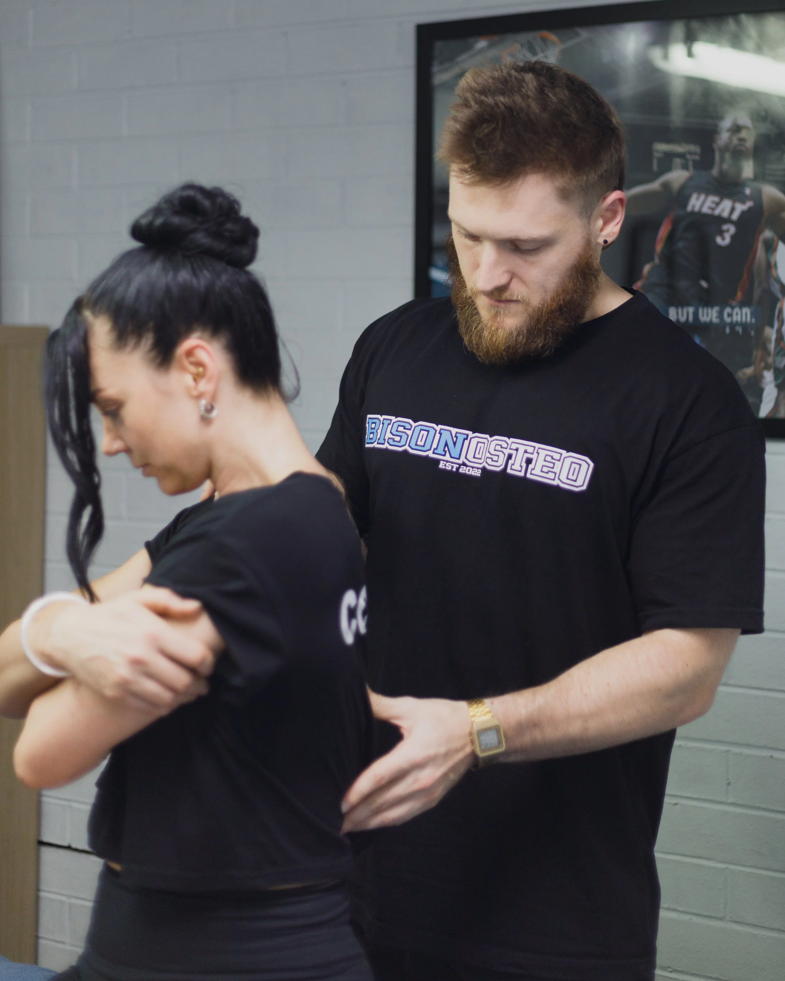 A man with a beard is helping a woman with a tattoo on her arm at a tattoo shop. The man is wearing a black t-shirt with the text 'BISONOSTEO' and a gold watch, while the woman has black hair in a bun and is wearing earrings.