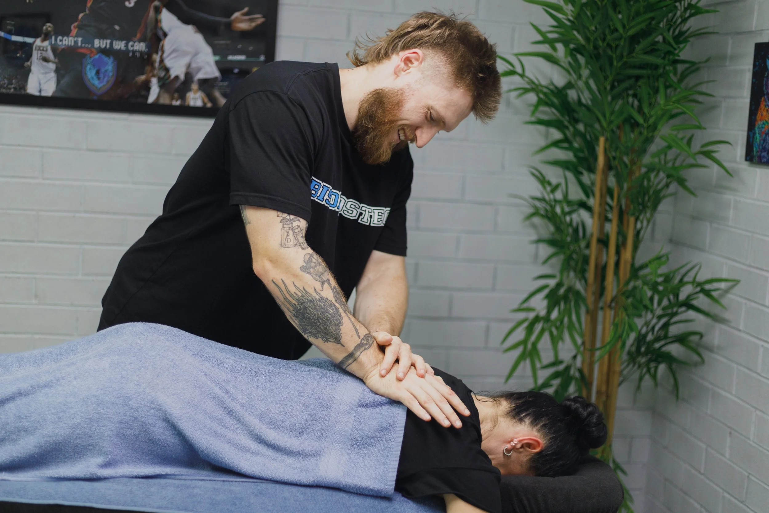 A man with tattoos and a beard gives a massage to a woman lying face down on a massage table, in a room with white brick walls, a tall green plant, and framed pictures on the wall.