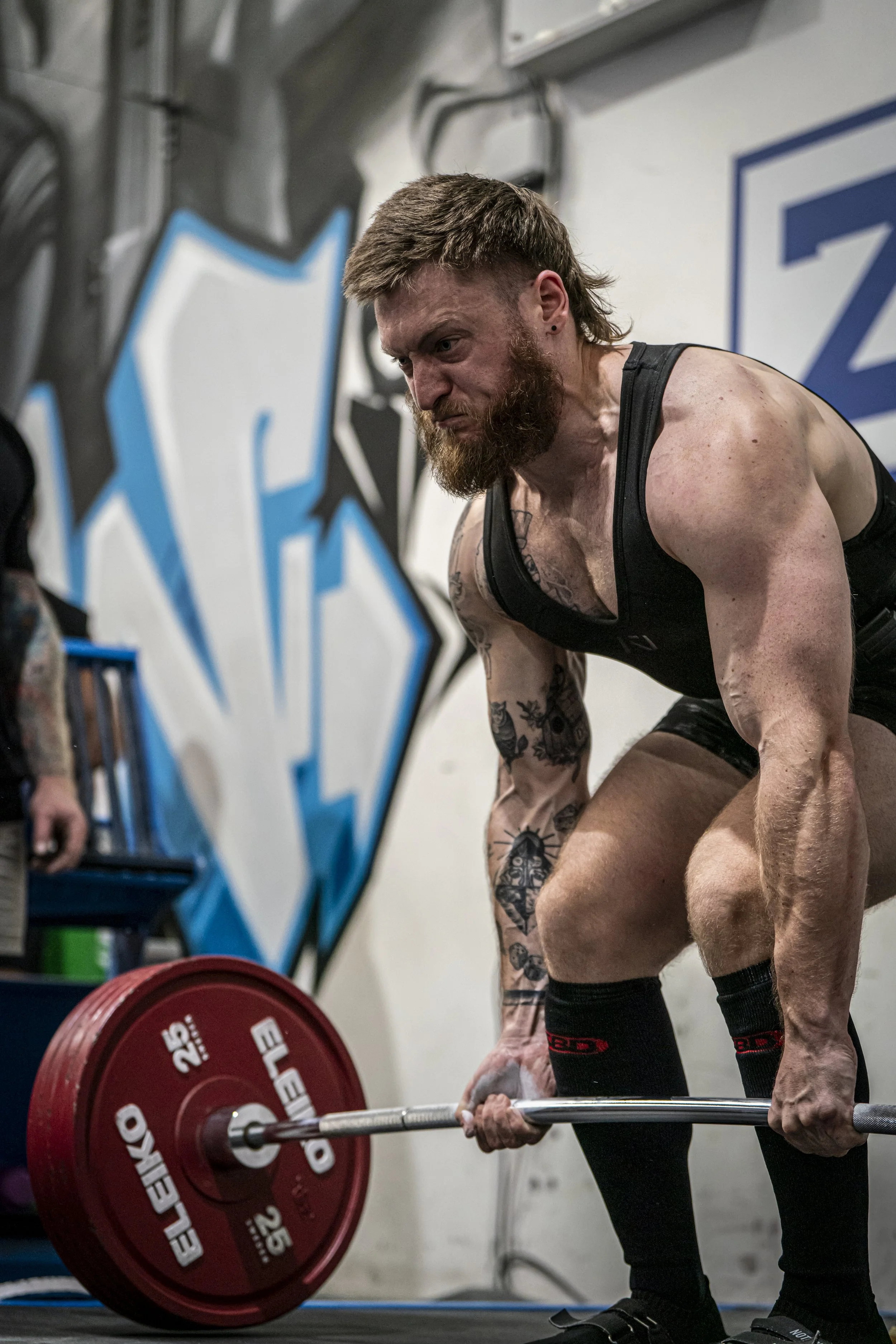 A muscular man with a beard lifting a red 25-pound weight plate during a workout in a gym with graffiti on the wall.