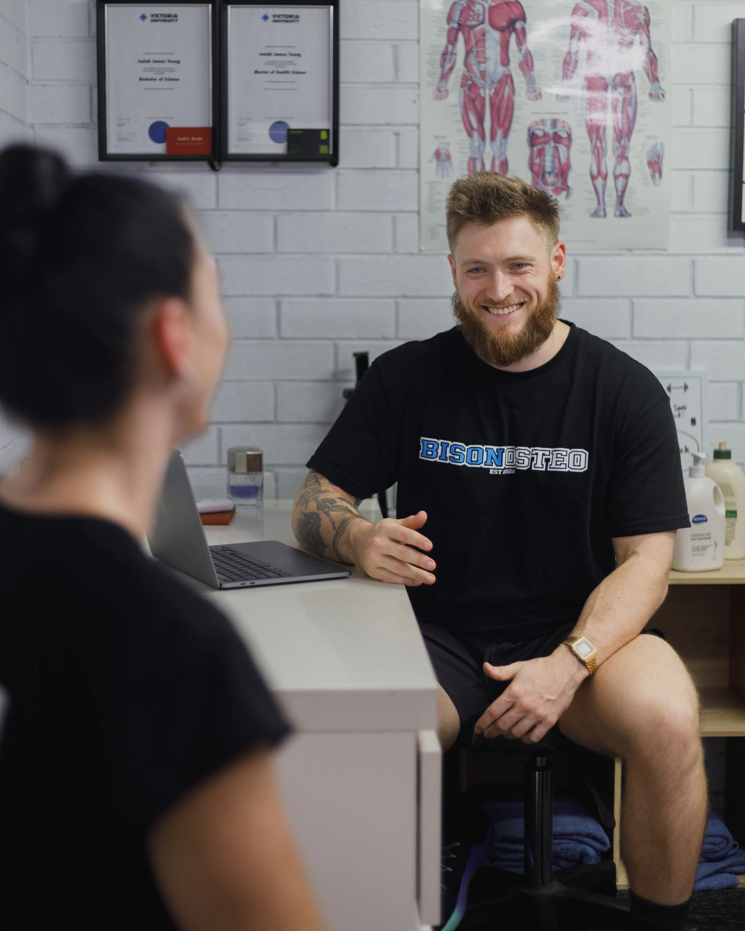 A man with a beard and tattooed arm smiling during a conversation with a woman in a medical setting, with anatomical charts and certificates on the wall.