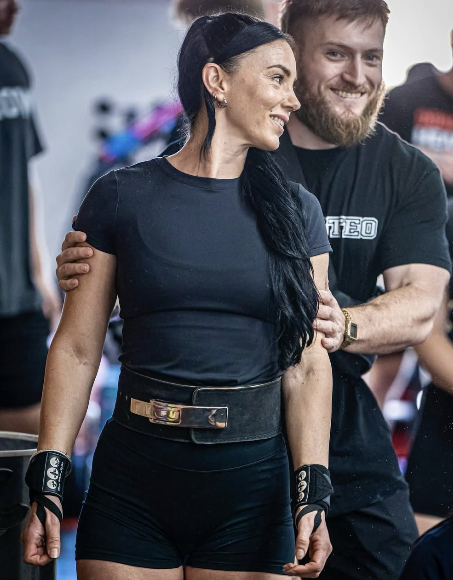 A woman with long black hair and a black headband smiling and standing with a man in a black T-shirt, who is holding her shoulder and smiling, in a social setting with other people in the background.
