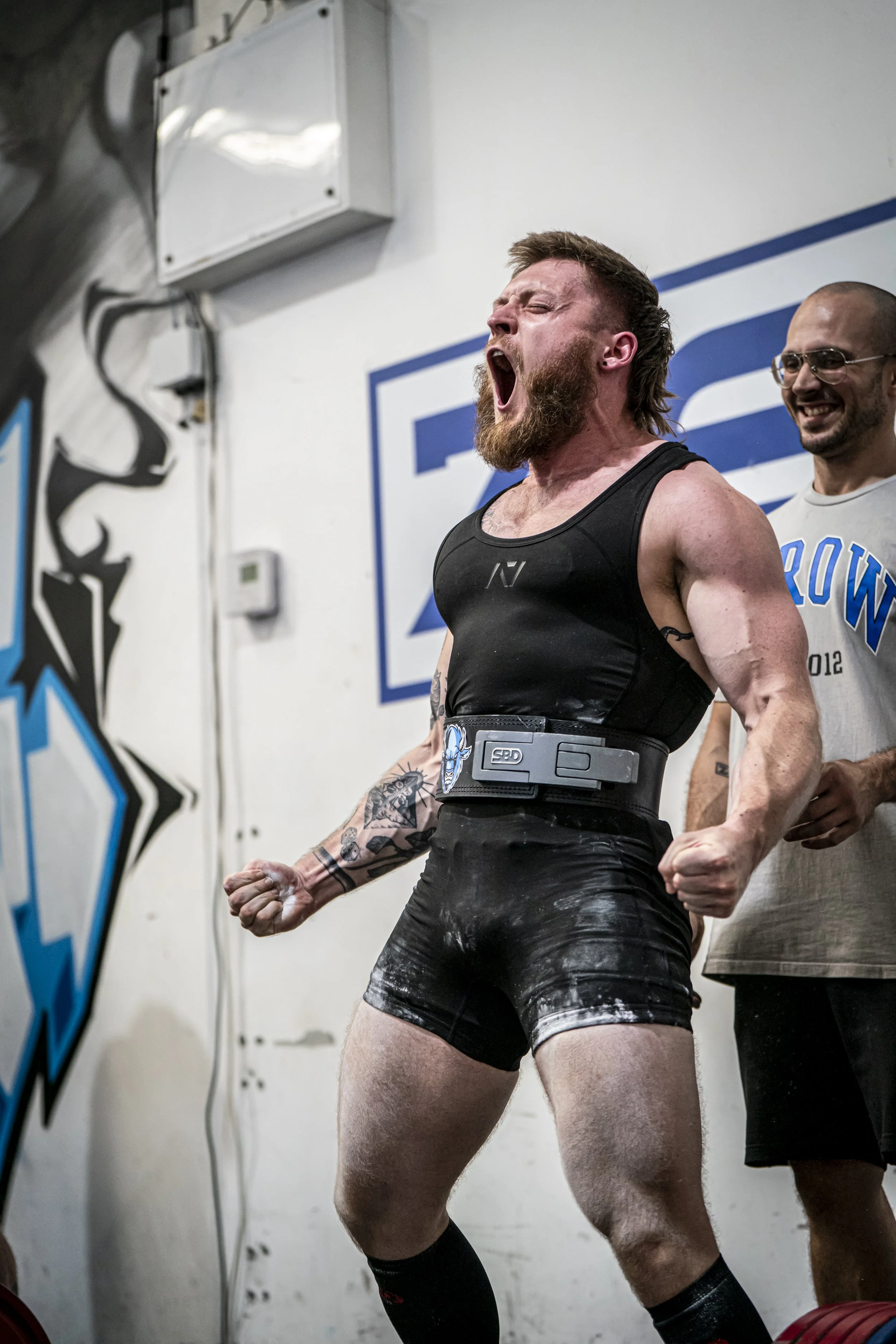 A muscular man with a beard wearing a black athletic outfit and a weightlifting belt, celebrating with a clenched fist and an open mouth, in a gym setting with another smiling man behind him.