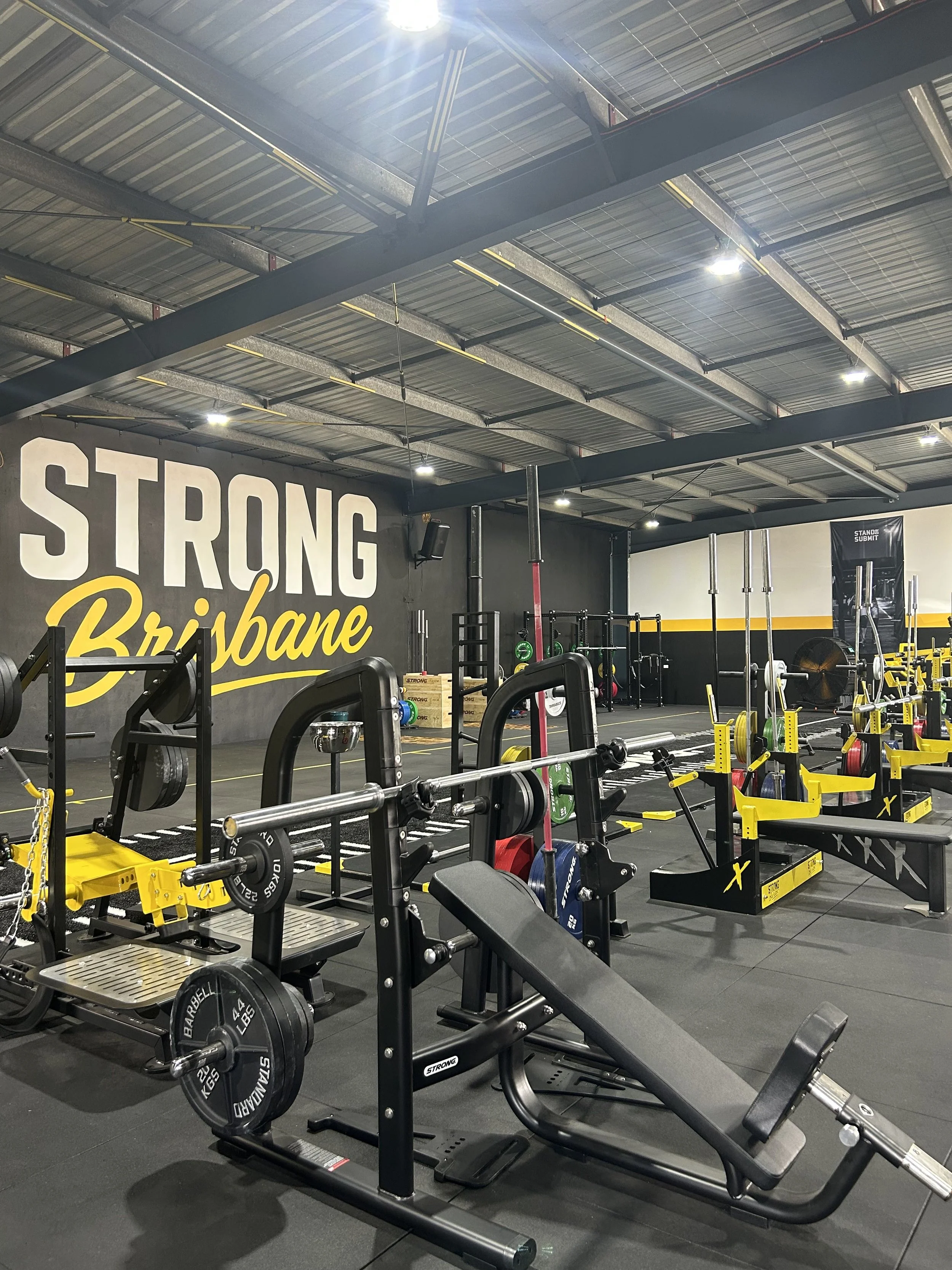 Indoor gym with various weightlifting equipment, including barbells, benches, and squat racks, with a black wall that says 'Strong Brisbane' in yellow and white lettering.
