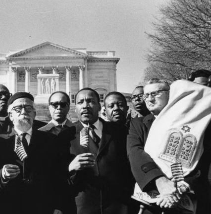 A group of men, including one holding a Torah scroll, standing in front of the Lincoln Memorial with the Parting of the Red Sea illustration on the scroll, during a day with clear skies.