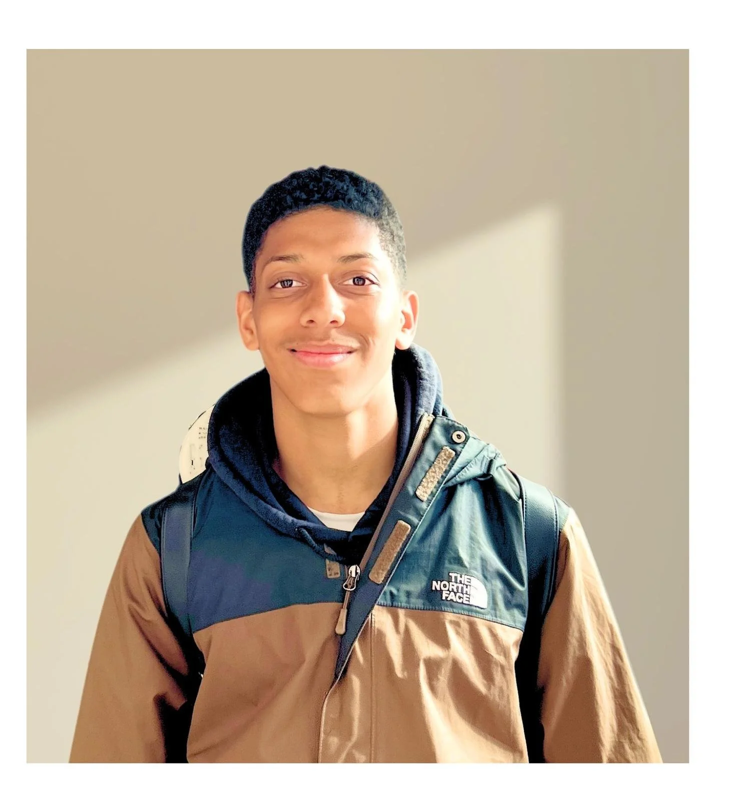 A young man with short black hair, smiling, wearing a brown and black North Face vest over a black hoodie, carrying a black backpack, standing indoors with a plain beige wall in the background.