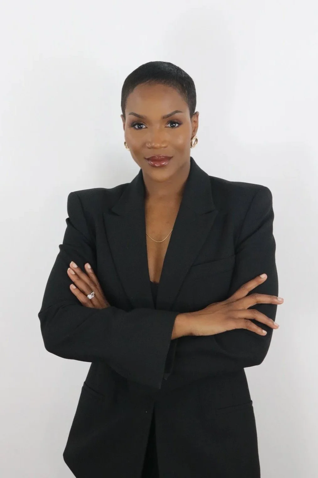 Portrait of an African American woman with short hair, wearing a black blazer, earrings, and a necklace, standing against a white background with arms crossed.