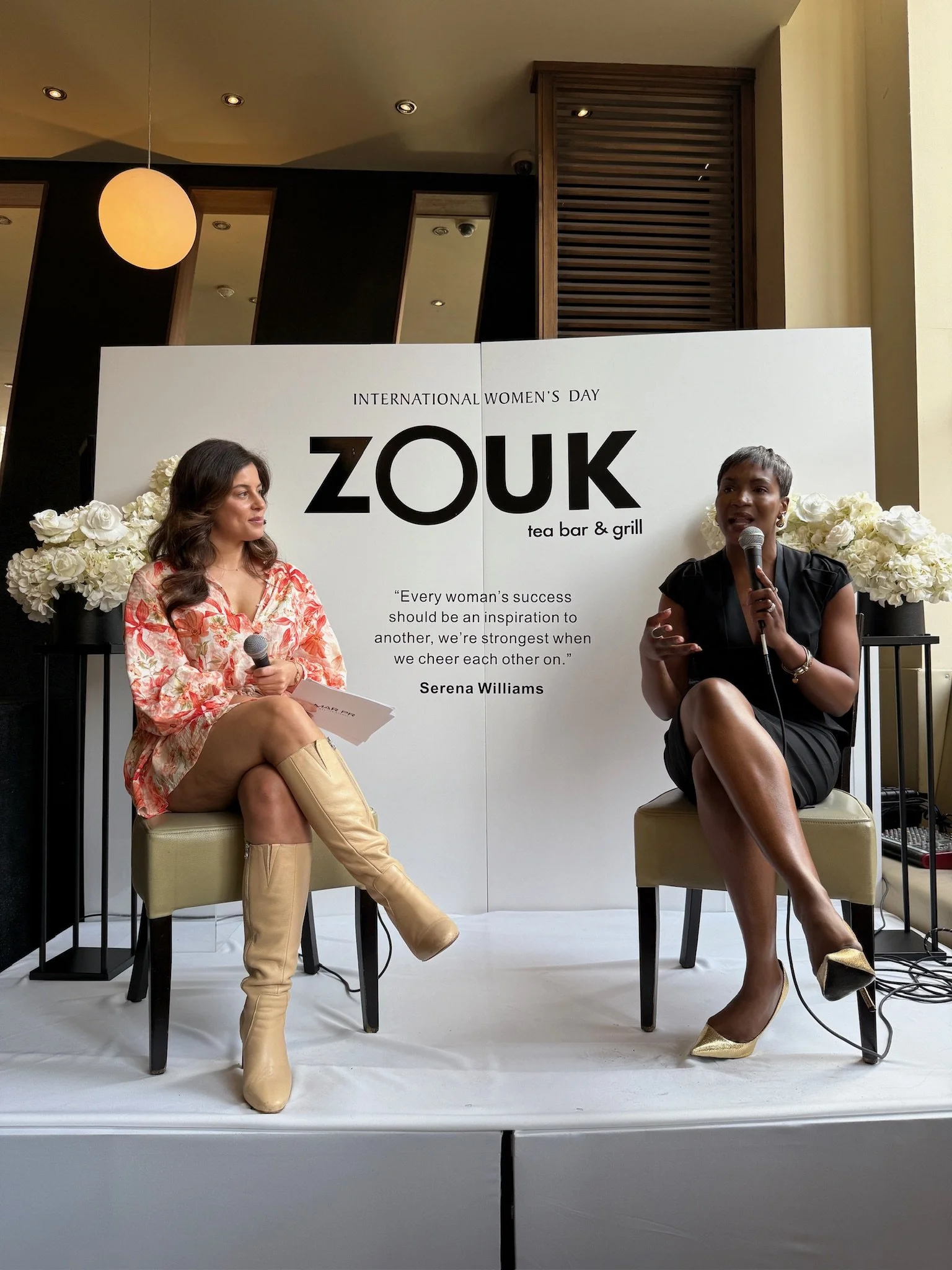 Two women sitting on chairs on a stage, participating in a discussion at Zouk tea bar & grill for International Women's Day. One woman, in a floral dress and beige knee-high boots, holds a microphone and some papers, while the other woman, in a black
