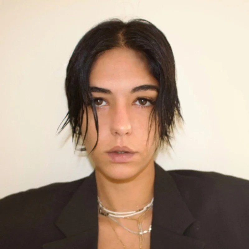 Portrait of a young woman with short black hair, wearing layered necklaces and a black blazer, standing against a plain light-colored background.