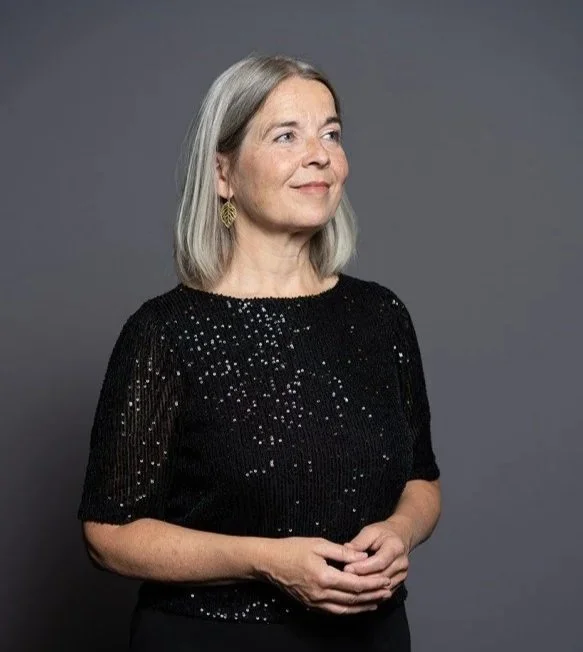 En ældre kvinde med gråt hår, der smiler let, iført en sort sweater med små glitrende elementer, stående mod en grå baggrund.