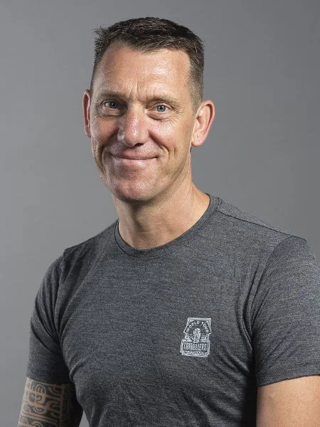 Ein Mann mit kurzen dunklen Haaren, grauem T-Shirt mit einem Logo, lächelnd vor grauem Hintergrund.