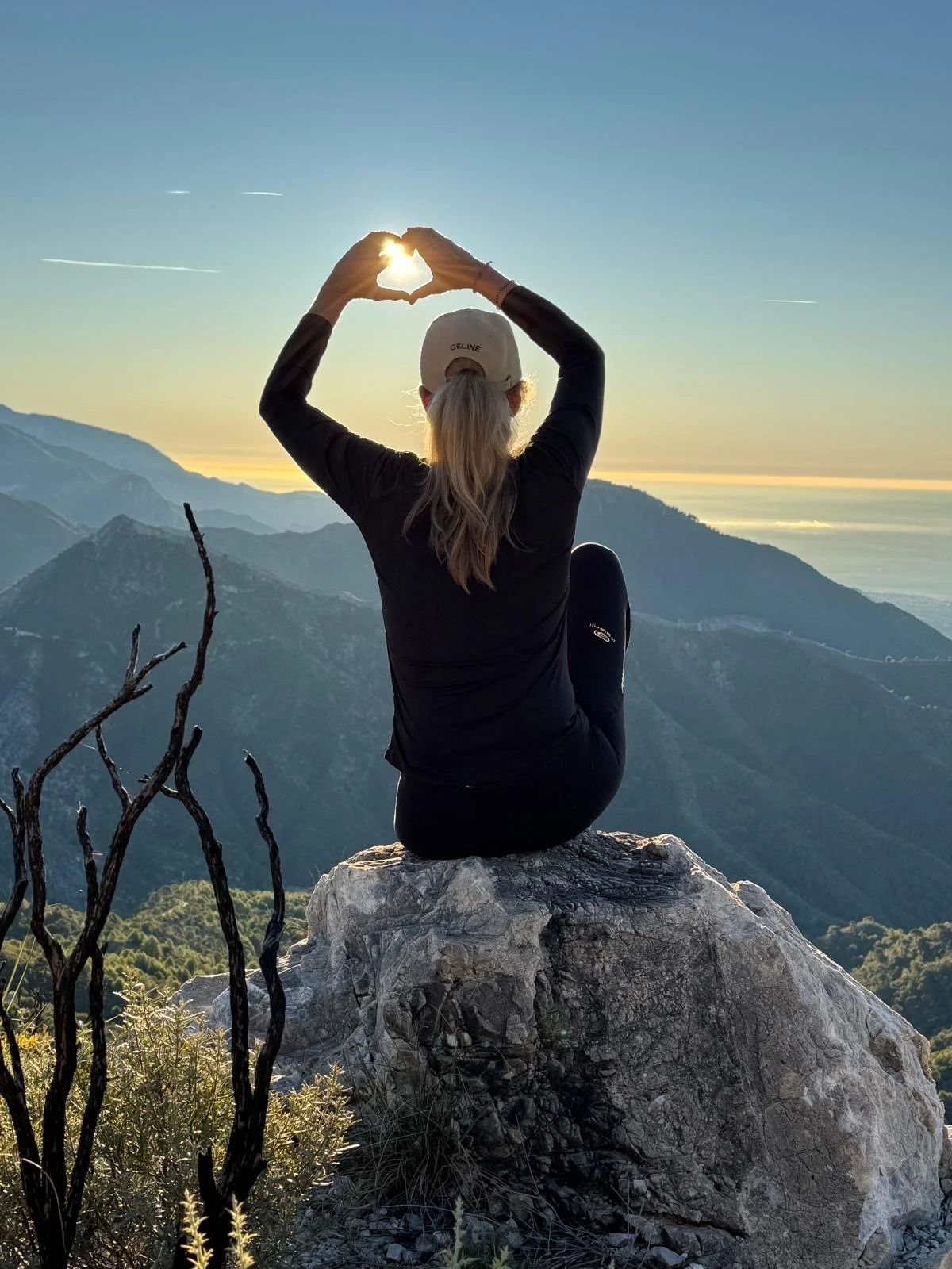 Persoon zit op een rots op een bergtop tijdens zonsopgang, maakt een hartvorm met haar handen, met een uitzicht op bergen en de oceaan op de achtergrond.