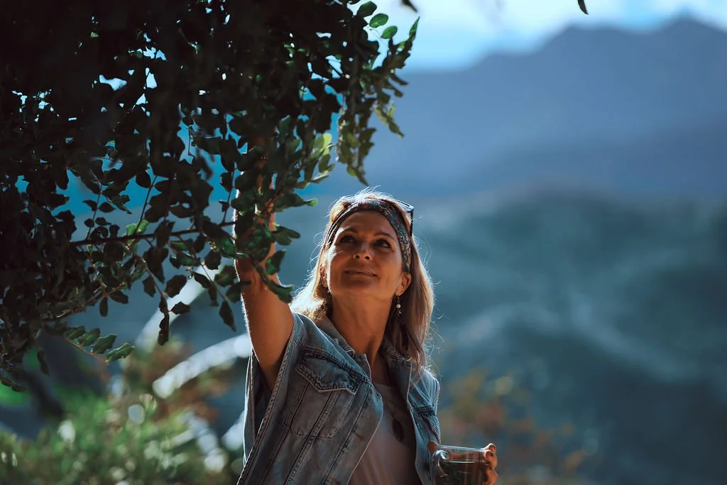 Een vrouw plukt bladeren van een boom in een natuurrijke omgeving met bergen op de achtergrond en een helderblauwe lucht.