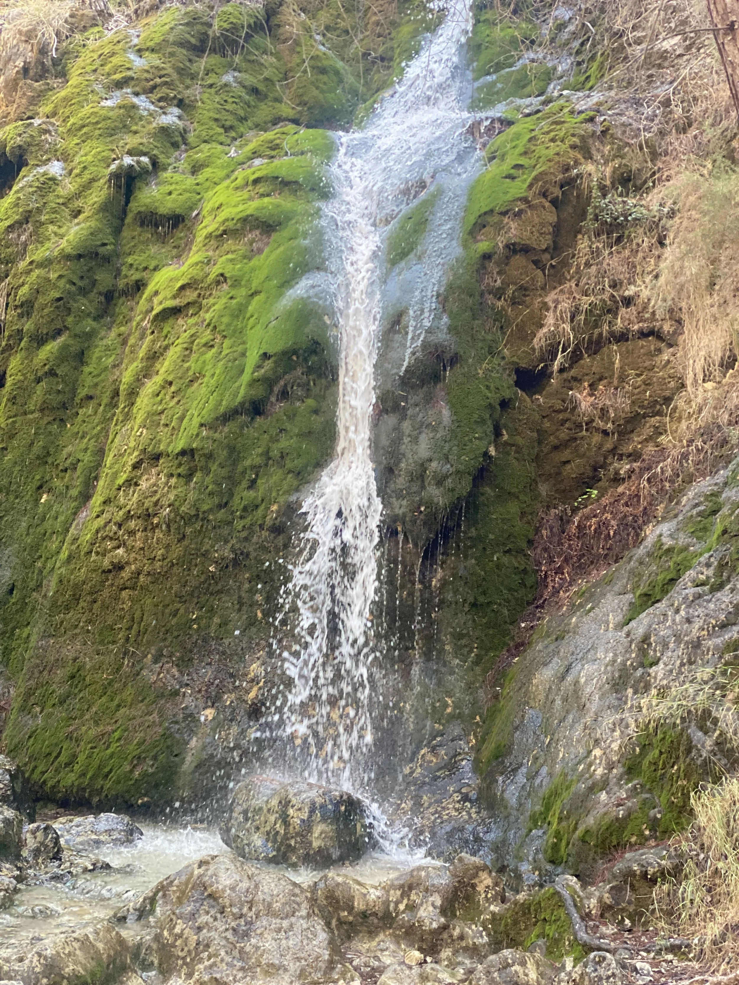 Een smal waterval die door een groen, mosbedekt rotslandschap naar beneden stroomt.