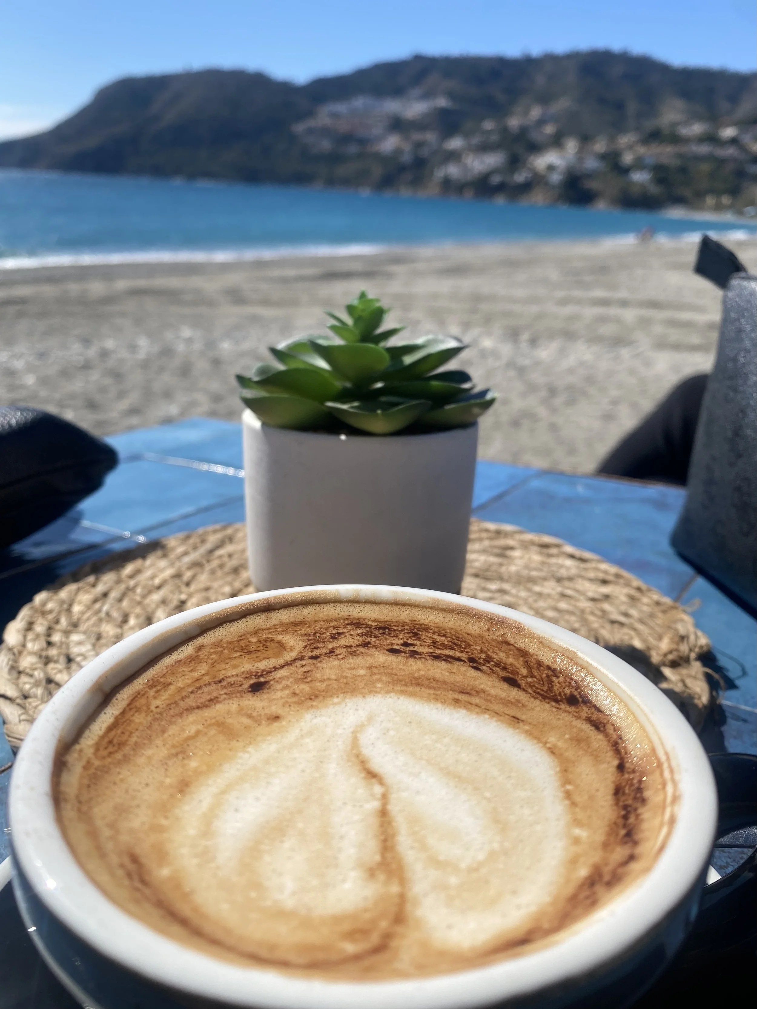 Een kop dampende koffie met crèmelaag en kaneel, op een tafel met een lapje stof en een drankje op de achtergrond, uitzicht op een strand met zee en bergen onder een blauwe lucht.