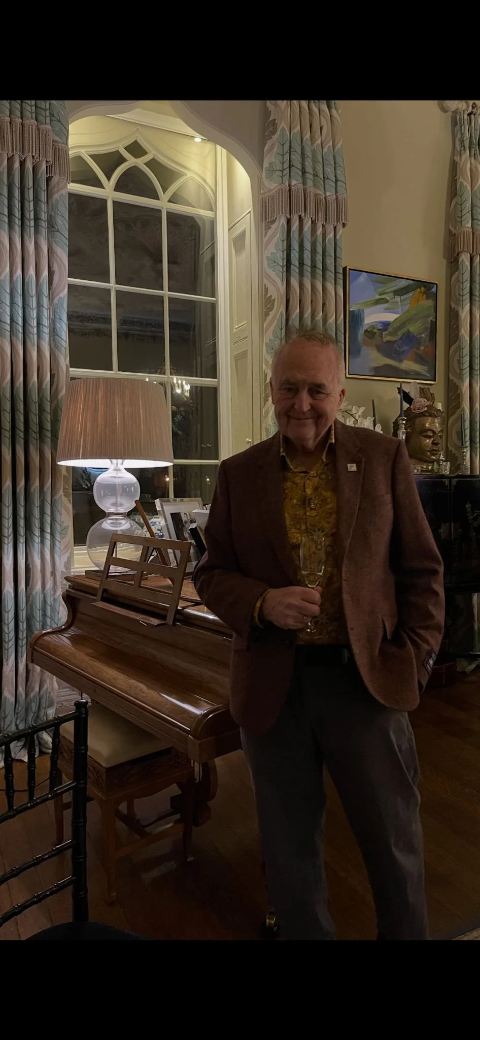 A man smiling and holding a champagne glass in a well-lit, elegant room with a large window, patterned curtains, a table lamp, framed photographs, a painting, and decorative sculptures.