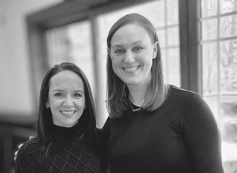 Two women smiling indoors with large windows in the background, one with shoulder-length dark hair and the other with shoulder-length light hair.