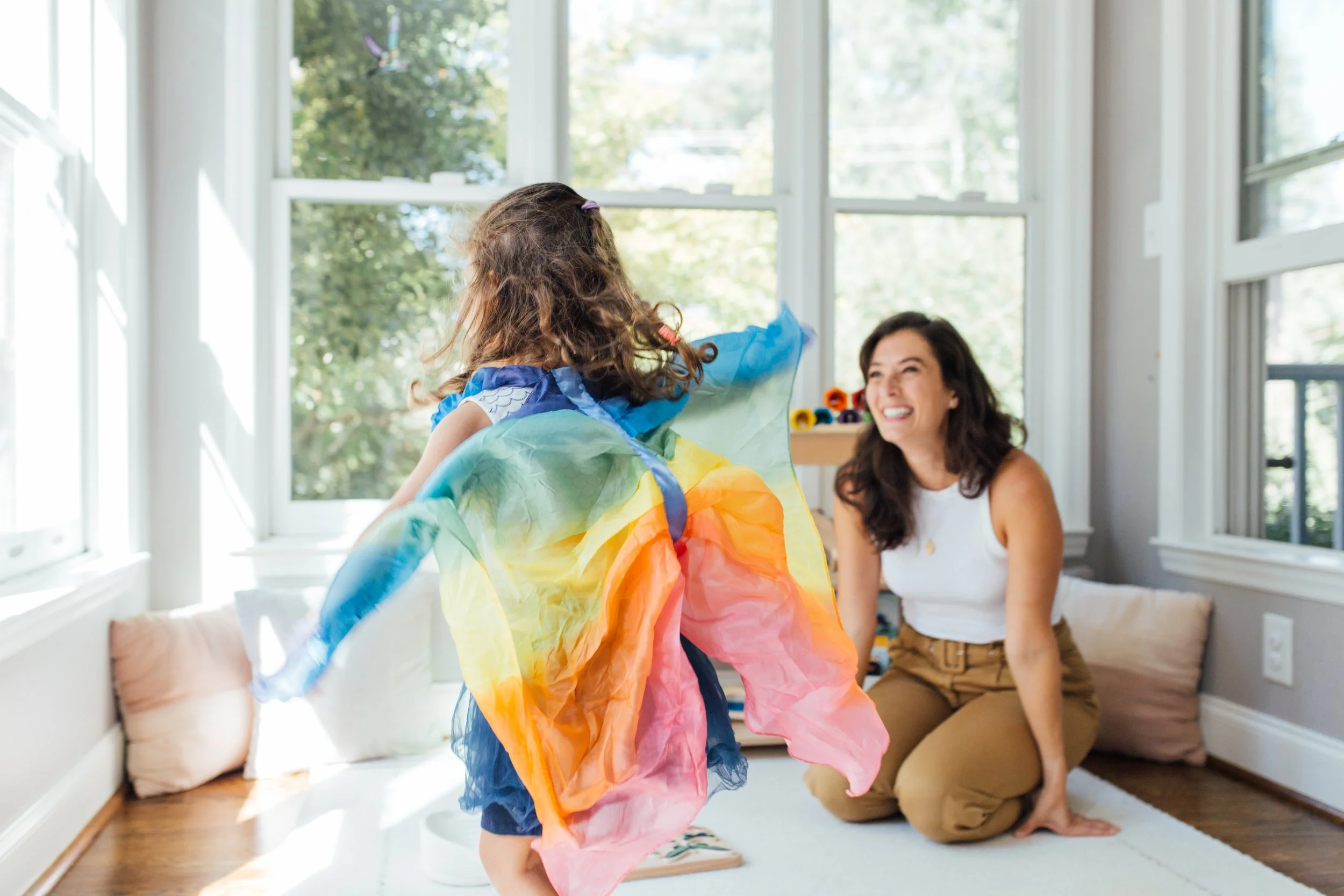 A young girl in a colorful princess dress is playing in a sunlit room with large windows, while an adult woman sits on the floor and smiles at her.