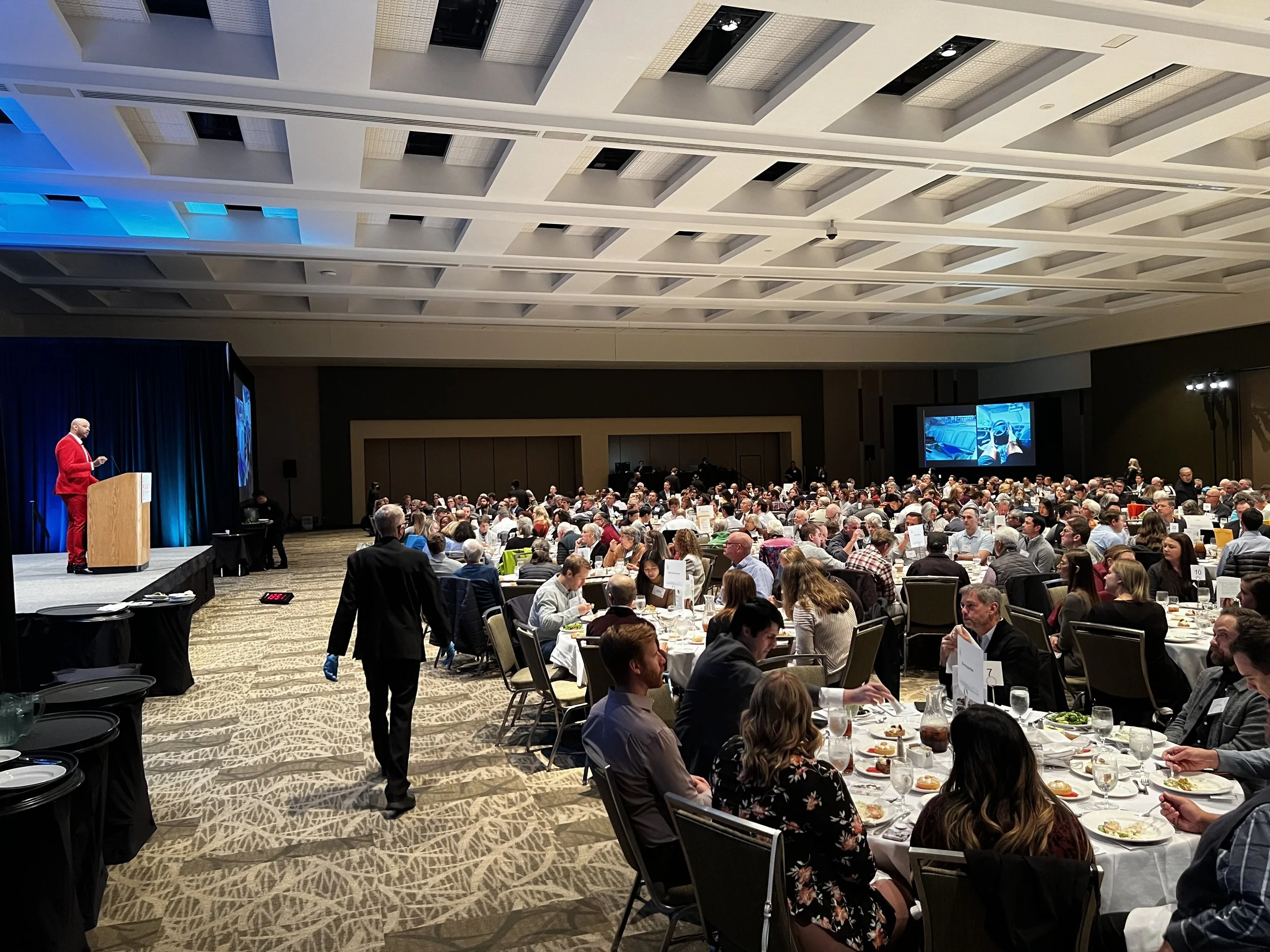 A large banquet hall filled with round tables of people attending a formal event. A speaker in a red suit stands at a podium on stage, addressing the audience. Two large screen TVs display images, and the room is well-lit with a high ceiling and patterned carpet.