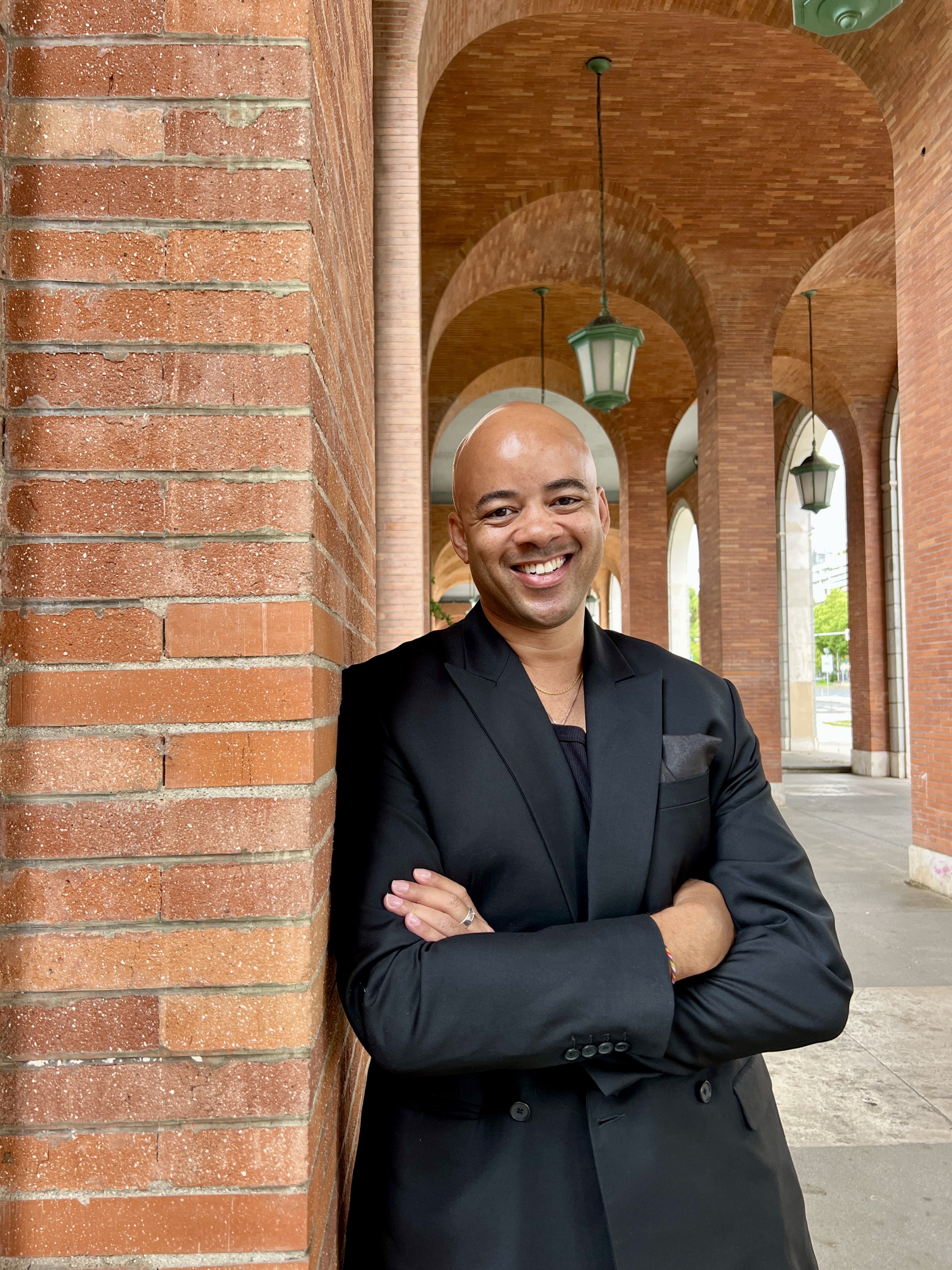 Photo of Ethan Ward in a black suit, arms crossed, leaning against a brick column under an arched walkway
