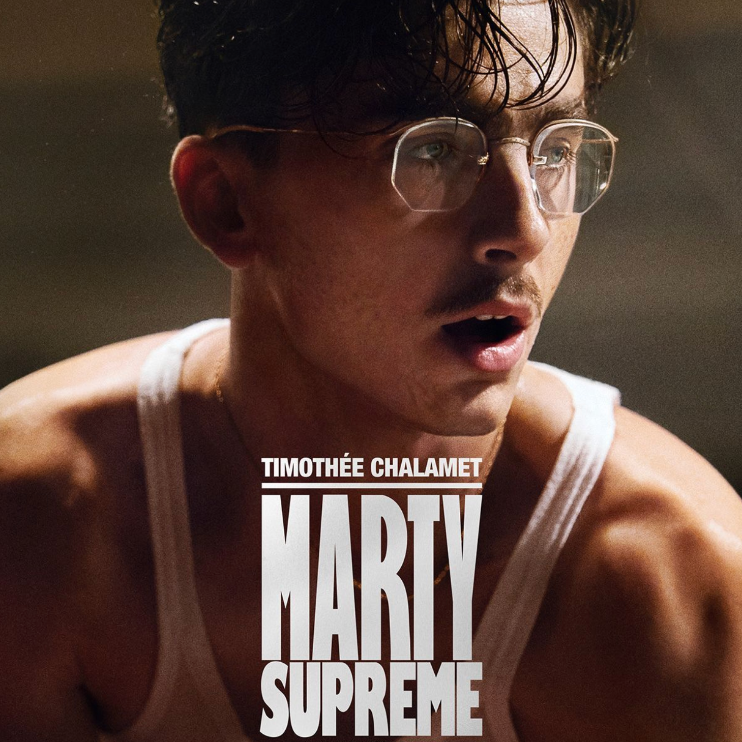 Close-up of a young man with dark hair, glasses, and a mustache, wearing a white tank top, looking surprised or focused.