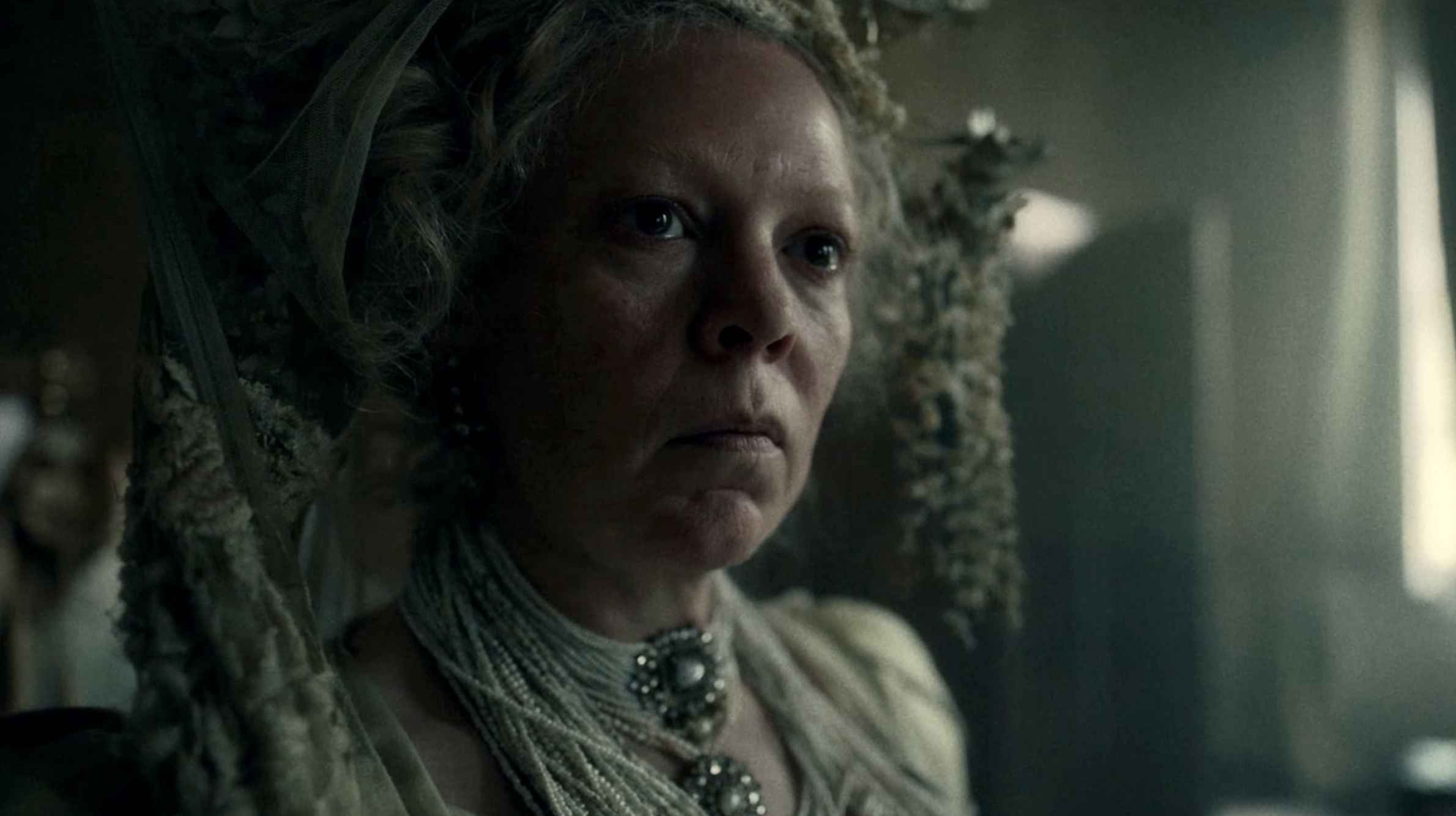 Close-up of a woman with a serious expression, wearing elaborate jewelry, and gazing out a window in a dimly lit room.