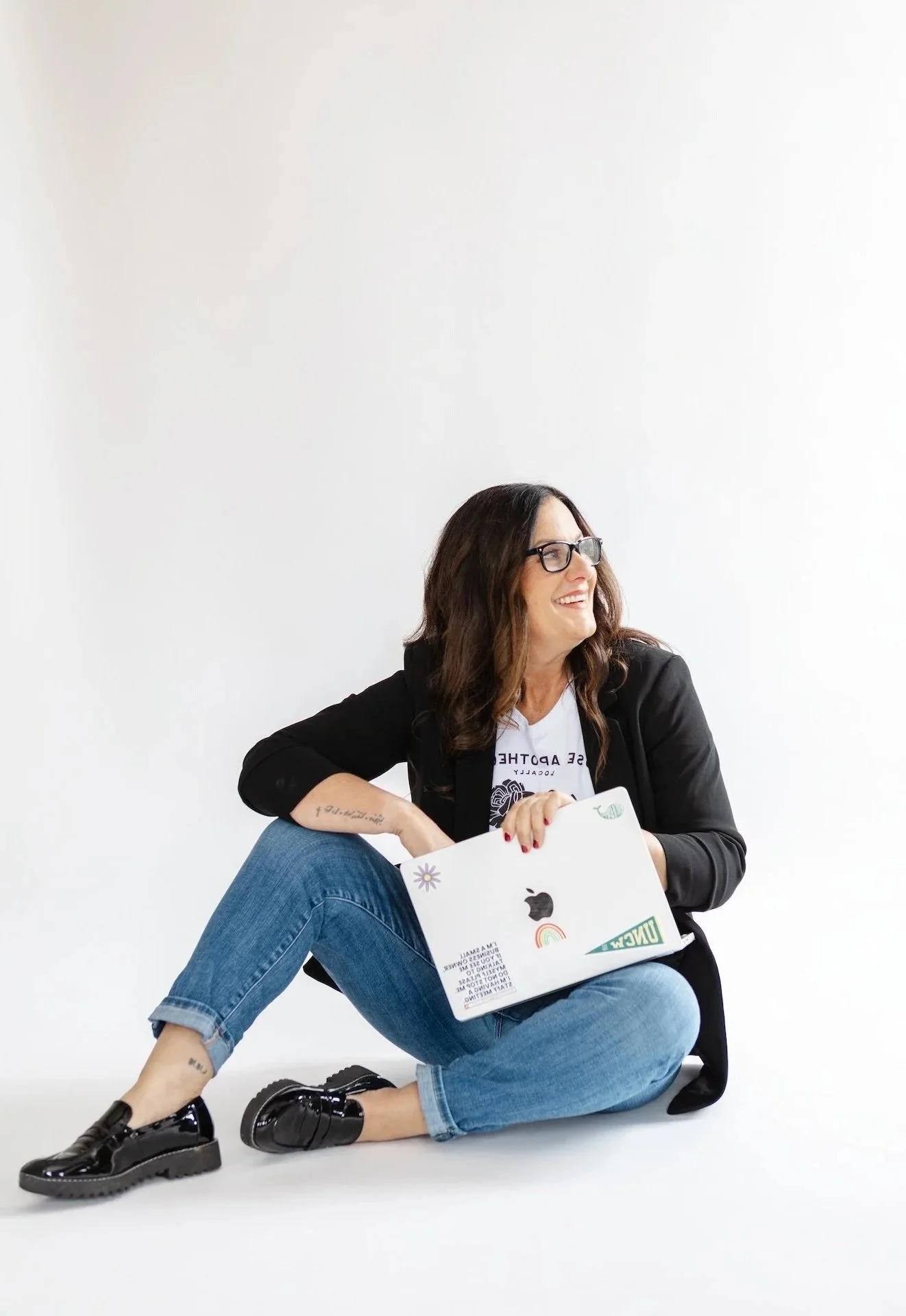 Woman sitting on the floor holding a MacBook, smiling, wearing glasses, black blazer, jeans, and black shoes, against a white background.