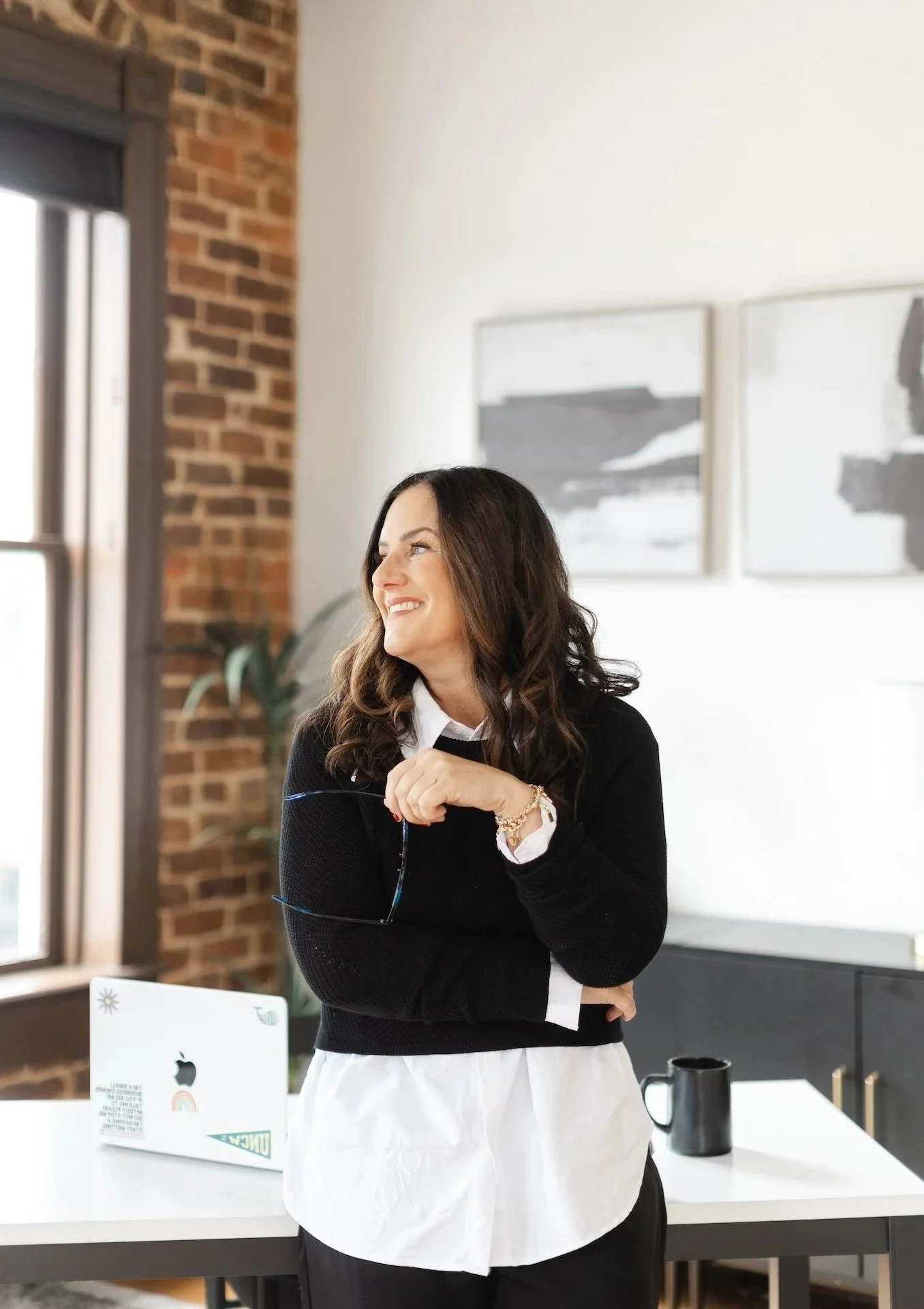 A woman with long brown hair, wearing a black sweater over a white shirt, is smiling and looking to the side in a modern office or studio with large windows, a white desk, a black mug, and abstract artwork on the wall.