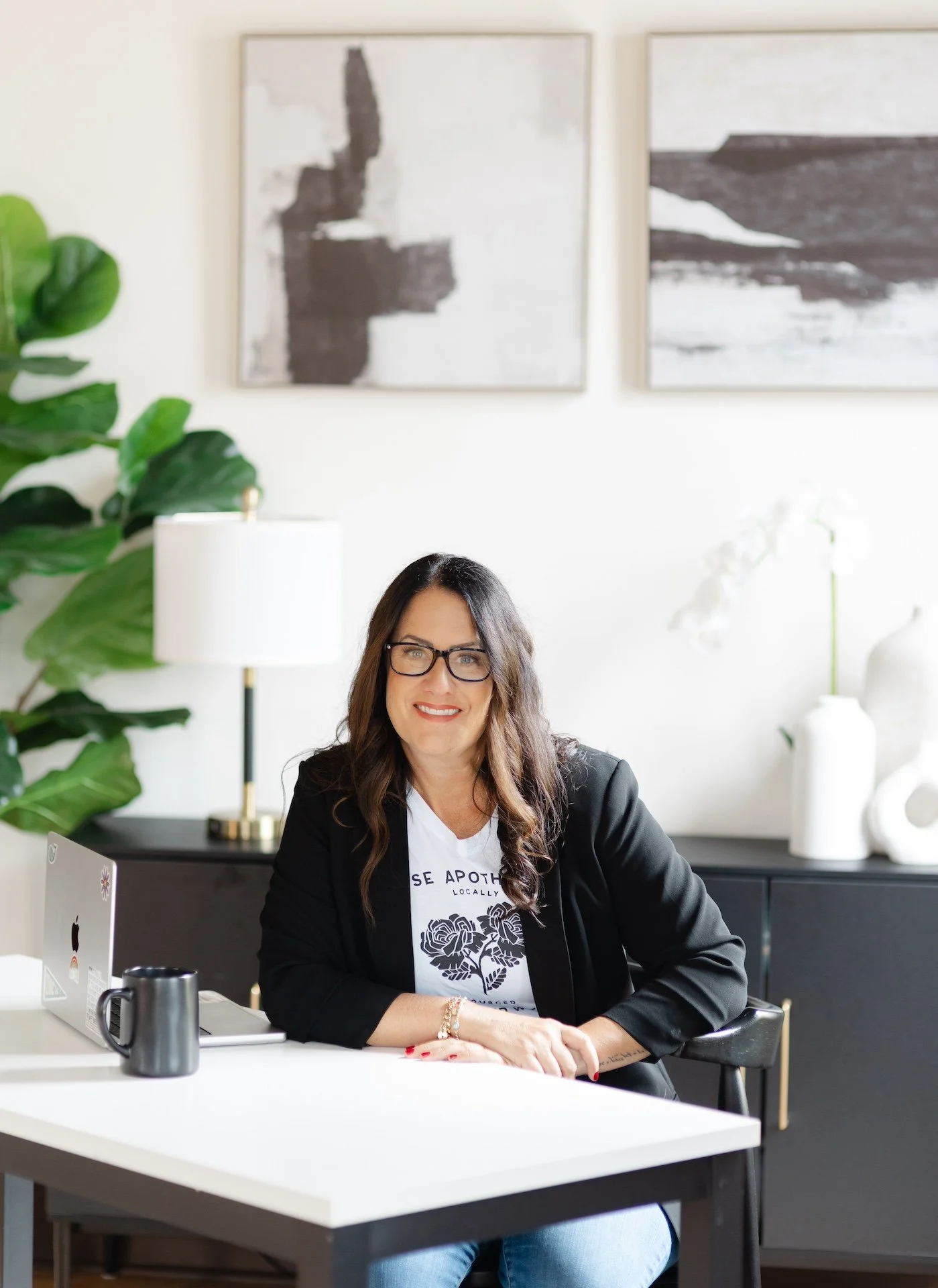 A woman with long dark hair, glasses, and a black blazer smiling while sitting at a white desk in a modern, minimalist office with abstract wall art and green plants.