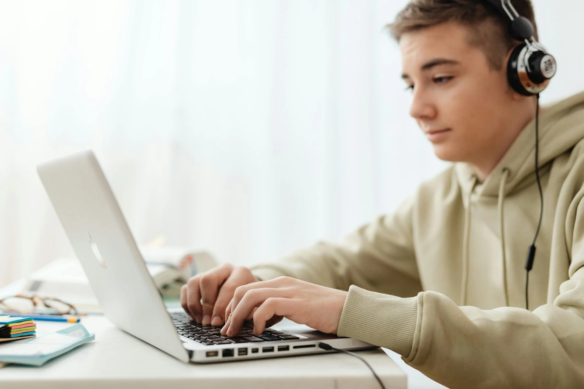 A young person with short brown hair wearing headphones and a beige hoodie working on a silver laptop at a white desk.