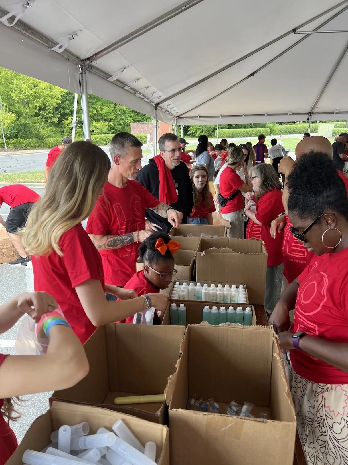 People working at a long table under a canopy during a community event, with boxes of bottles and tubes.