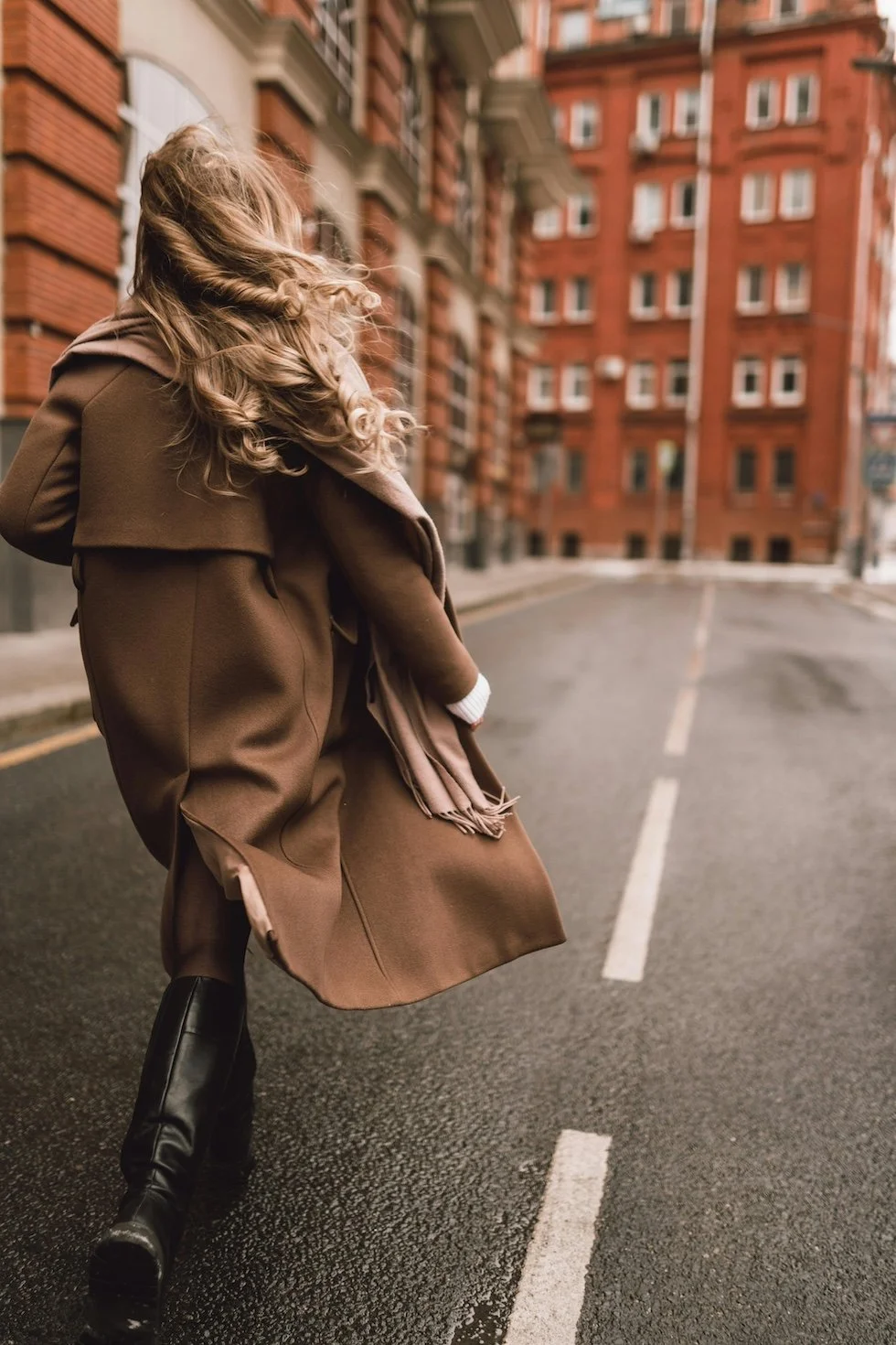 A woman walking on a city street with tall red brick buildings in the background, wearing a brown coat and black boots.