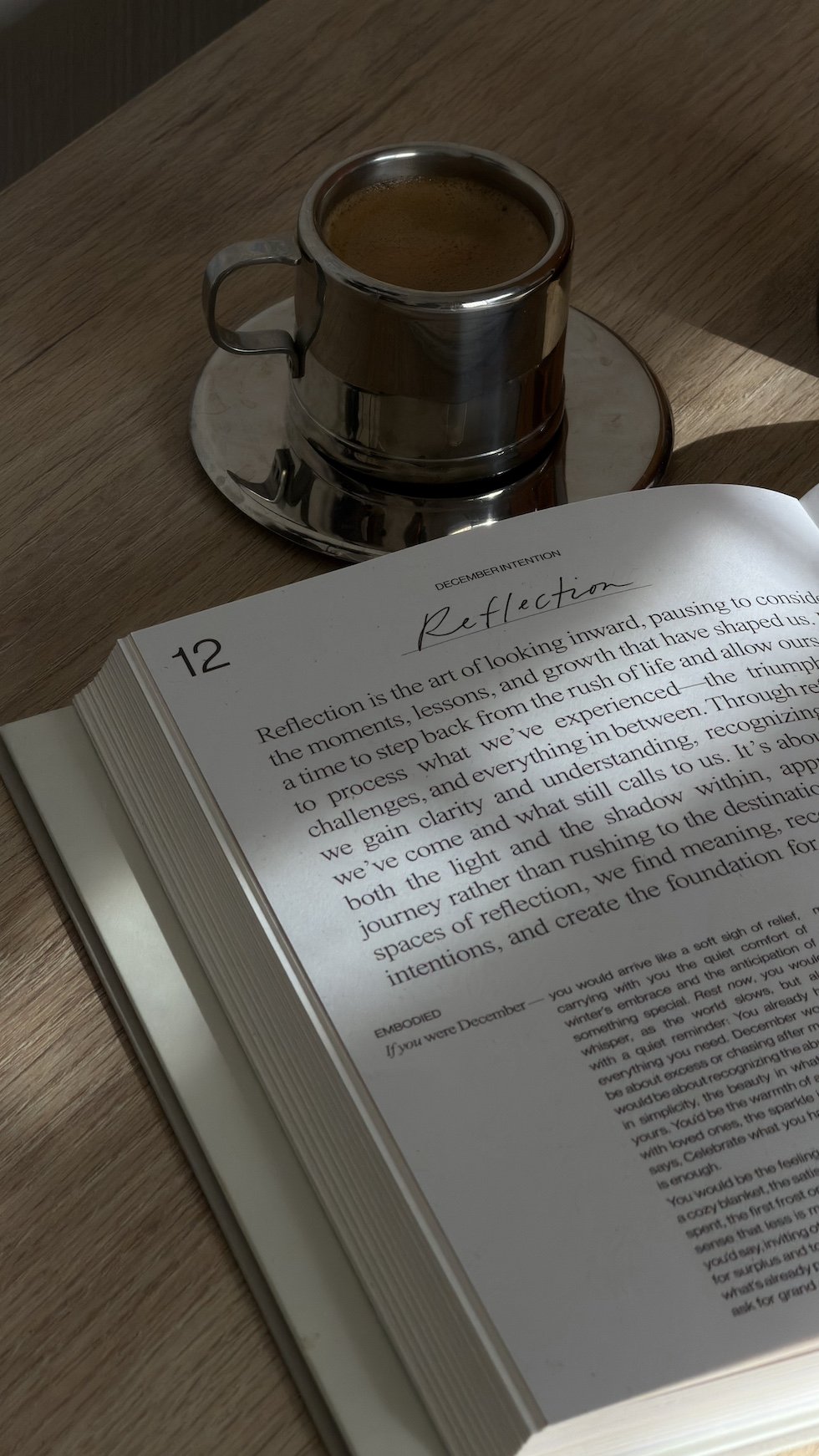 A glass cup filled with coffee on a saucer next to an open book with the title 'Reflection' on a wooden table.