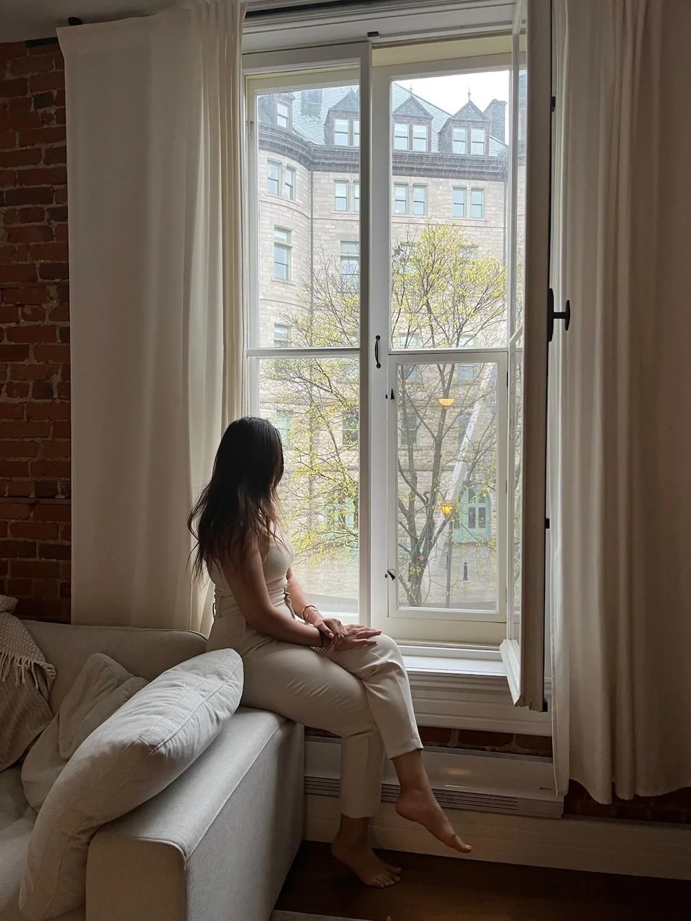 Woman sitting on a sofa near a large window, looking outside at a tree and a building.