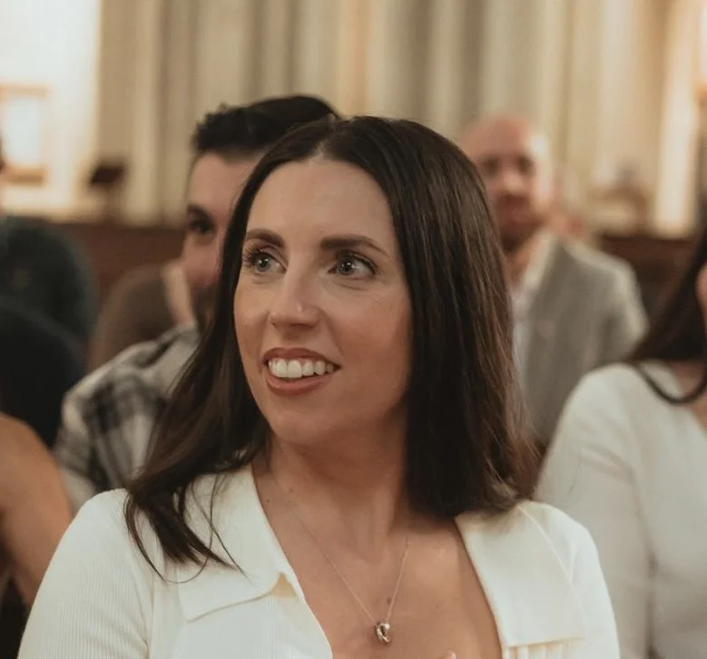 A woman with shoulder-length brown hair and a white top smiling at an indoor event, with other people in the background.