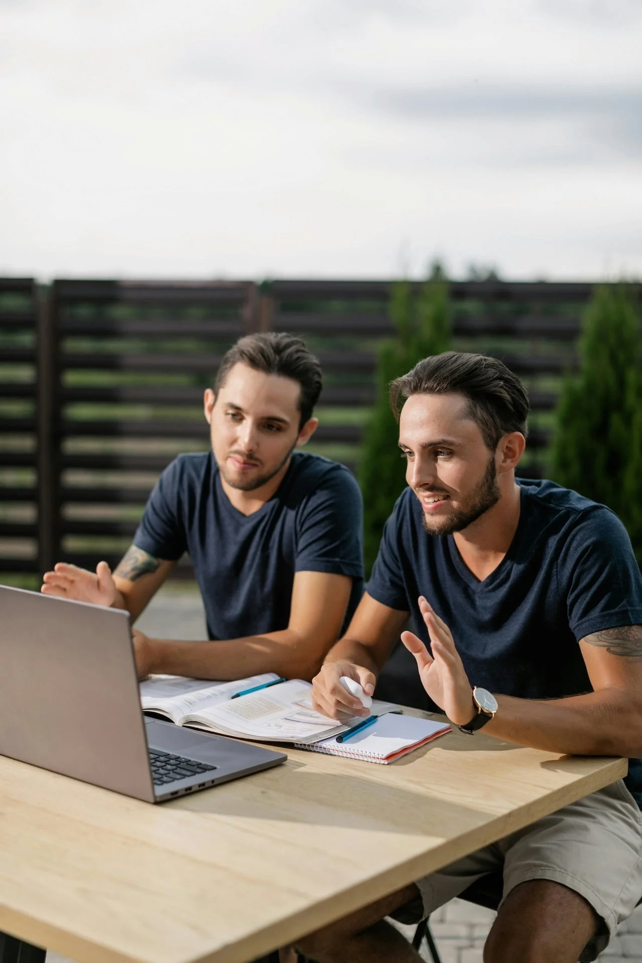 Two men seated at a table outdoors working on a laptop and discussing, with open books and notebooks in front of them.