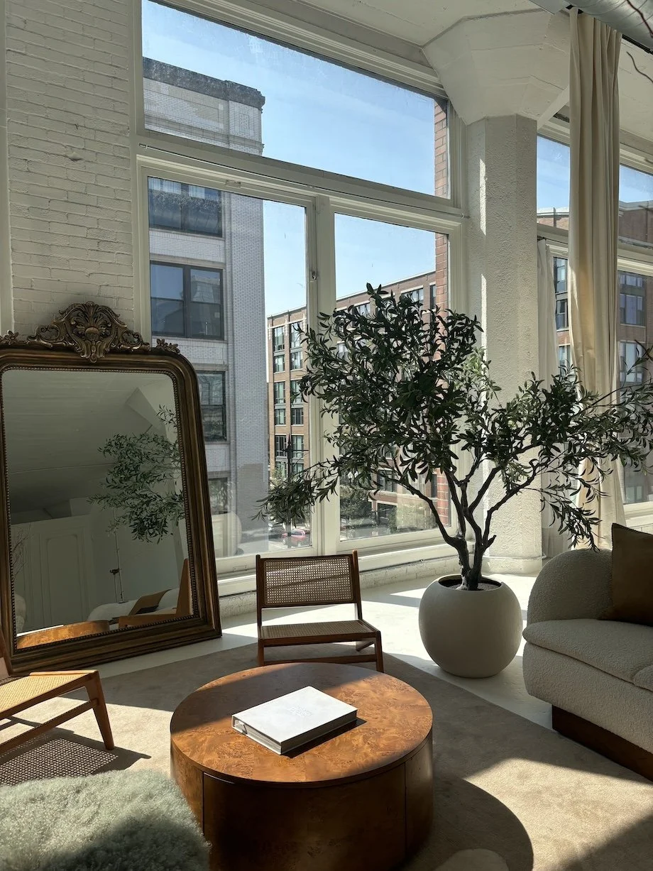 Living room with large windows, a potted plant, a mirror, a sofa, a wooden coffee table, and chairs.