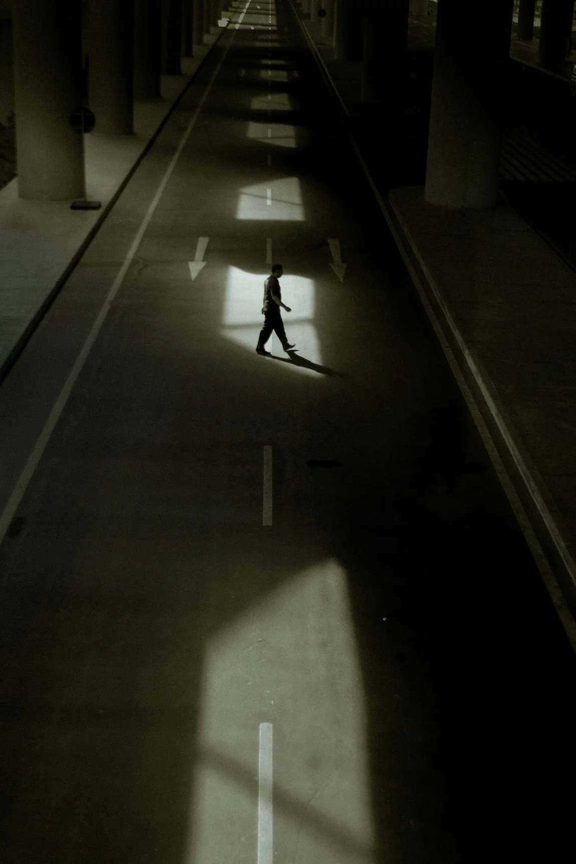 A solitary person walking on a dark empty street with sunlight casting shadows through openings in an urban setting.